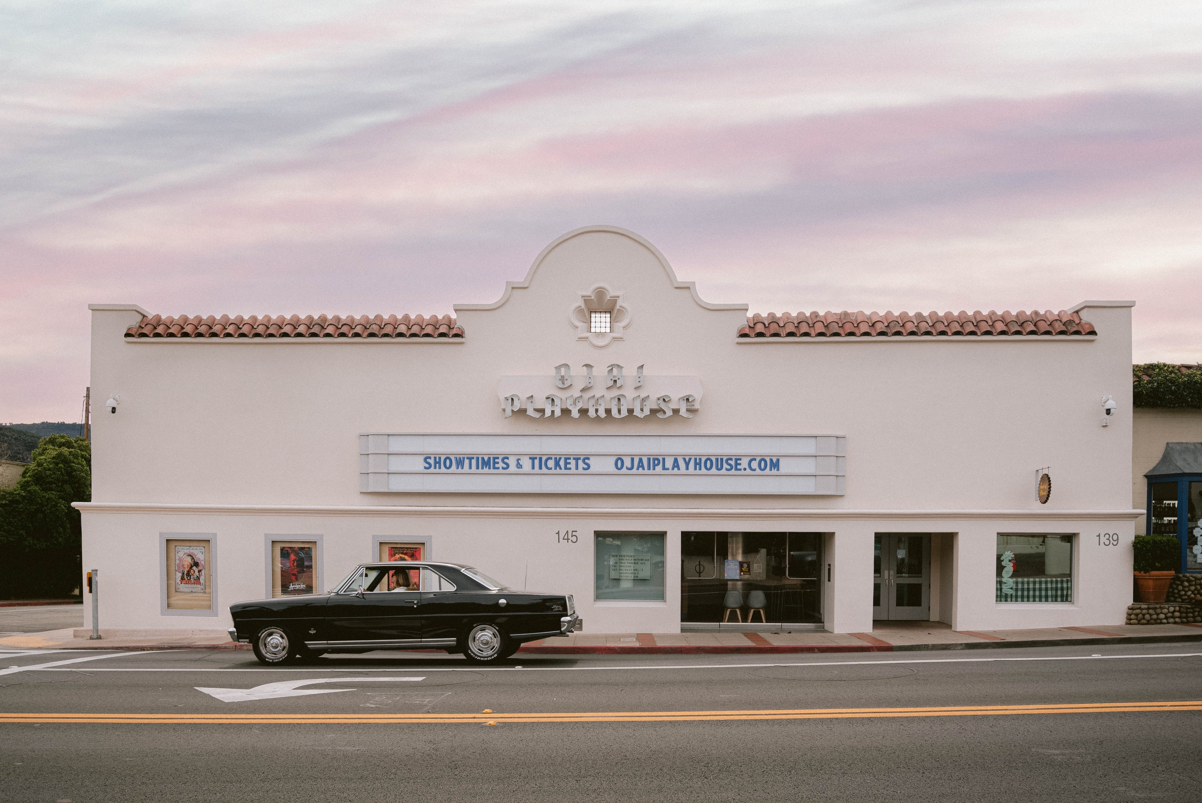 White building with a marquee and a black car parked in front, under a pink and purple sky.