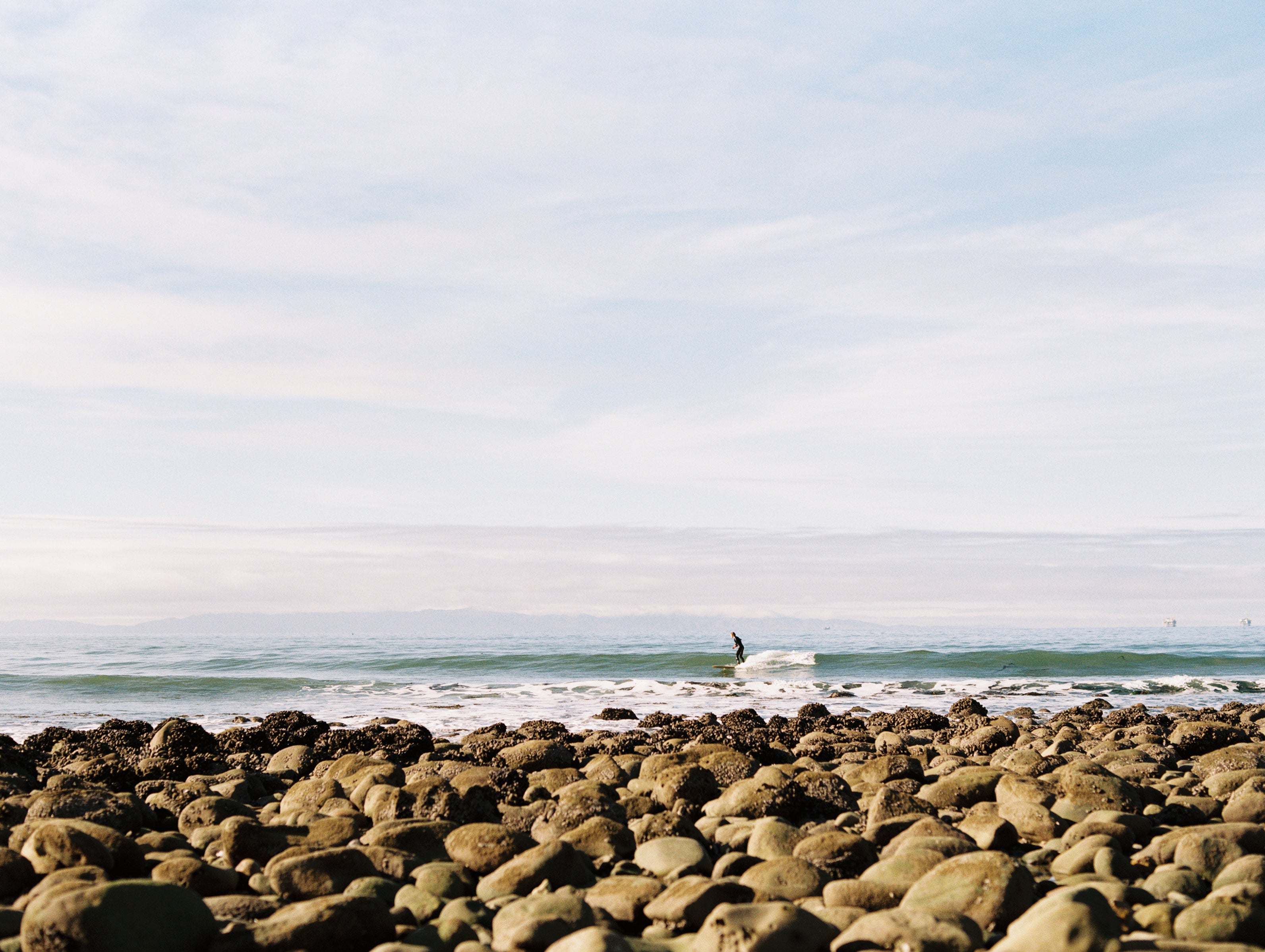 Person on a surfboard in the ocean with a rocky shore and clear sky.